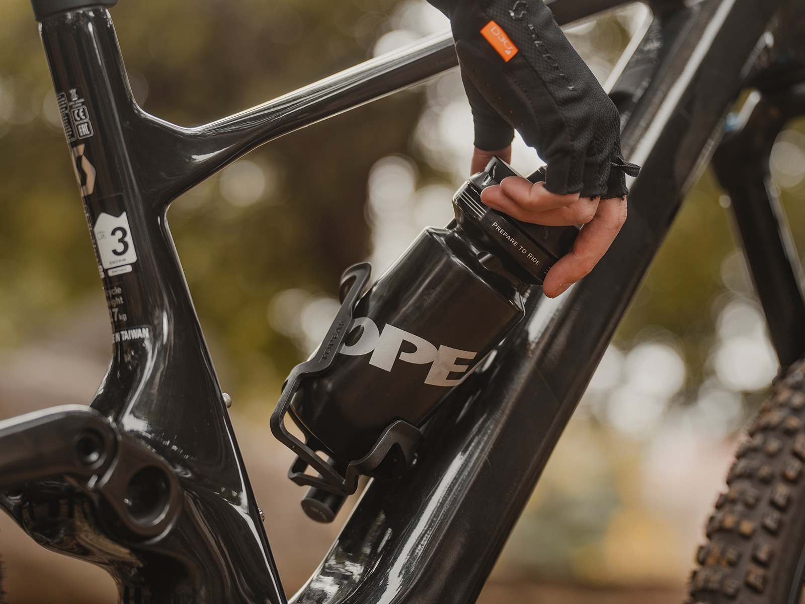 Hand eines Fahrers, der eine schwarze Topeak-Trinkflasche aus einem seitlich öffnenden Topeak-Flaschenhalter an einem Mountainbike-Rahmen herausnimmt.