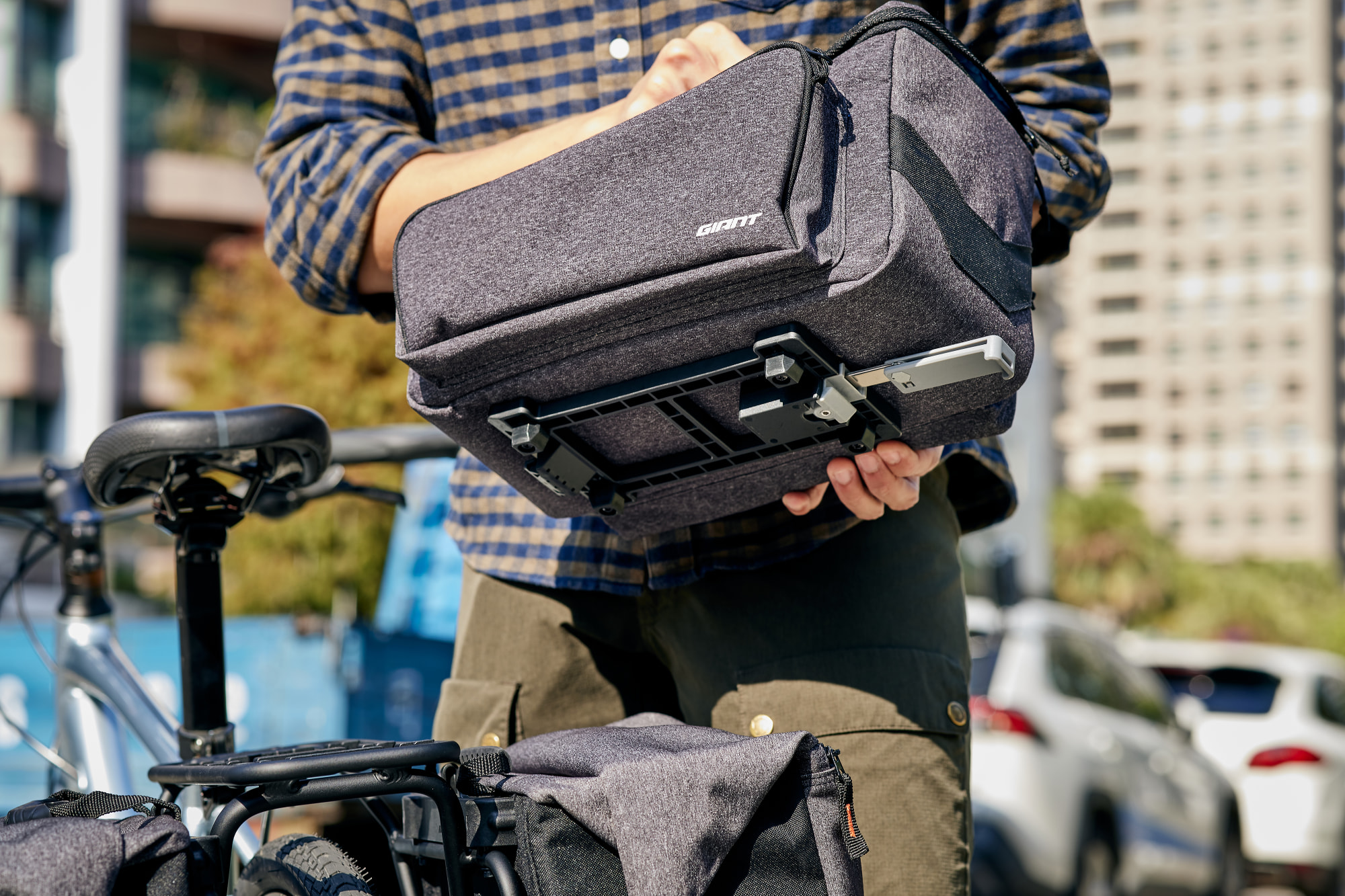 Person montiert eine Giant Gepäckträgertasche auf einem Fahrrad mit Rack-It Lite Gepäckträger, urbaner Hintergrund mit Gebäuden.