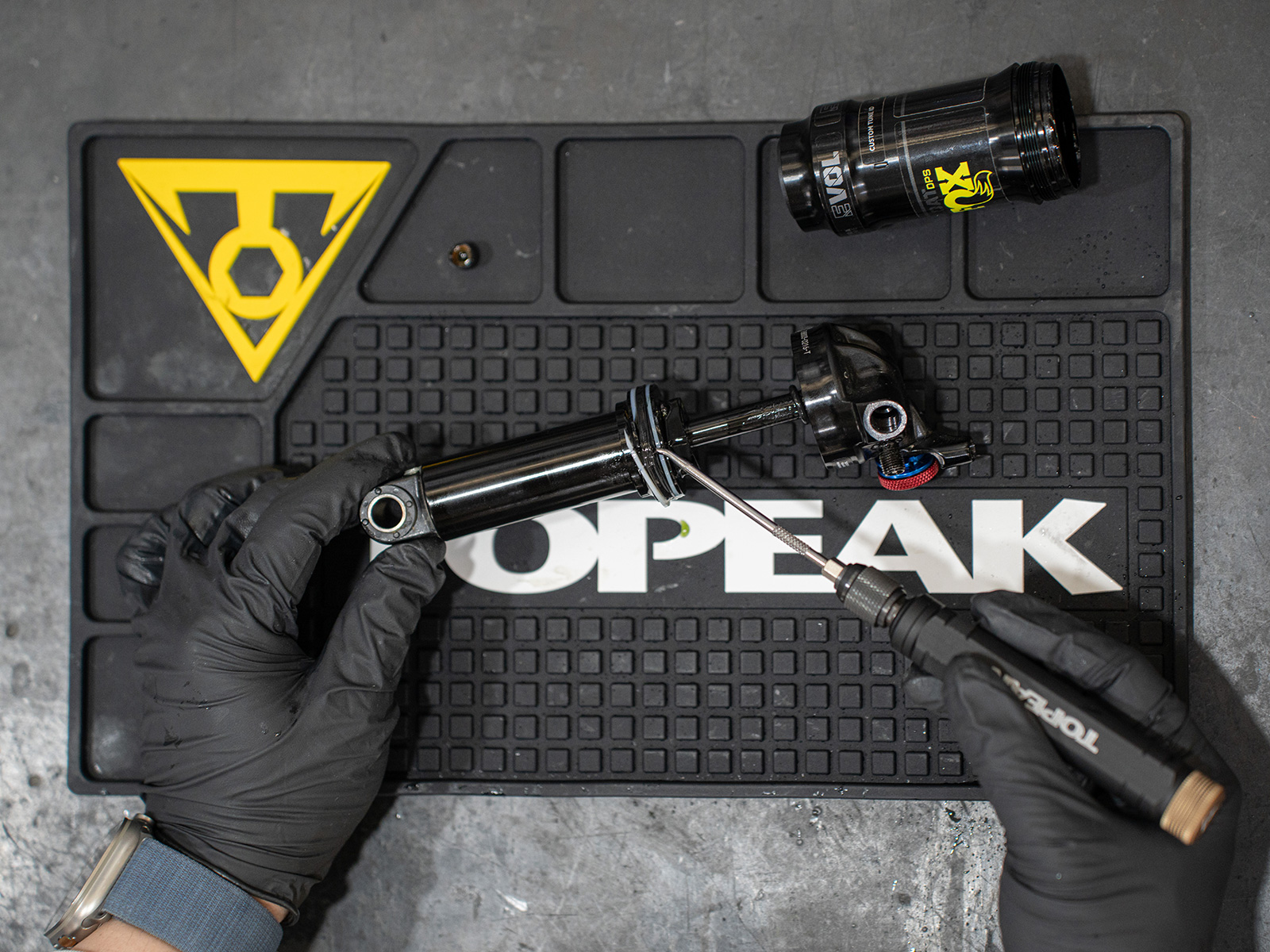 Topeak Silikon-Werkbankauflage im Einsatz – schwarze Matte mit gelbem Topeak-Logo und Unterteilungen, darauf Fahrrad-Dämpfer und Werkzeug bei Wartungsarbeiten.