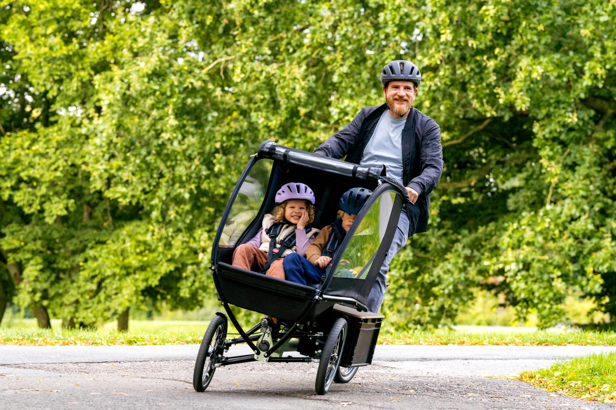 Ein Erwachsener fährt ein Chike e‑Kids Lastenrad mit zwei Kindern in der überdachten Frontkabine einen Weg im Park entlang. Die Umgebung ist von grünen Bäumen geprägt.
