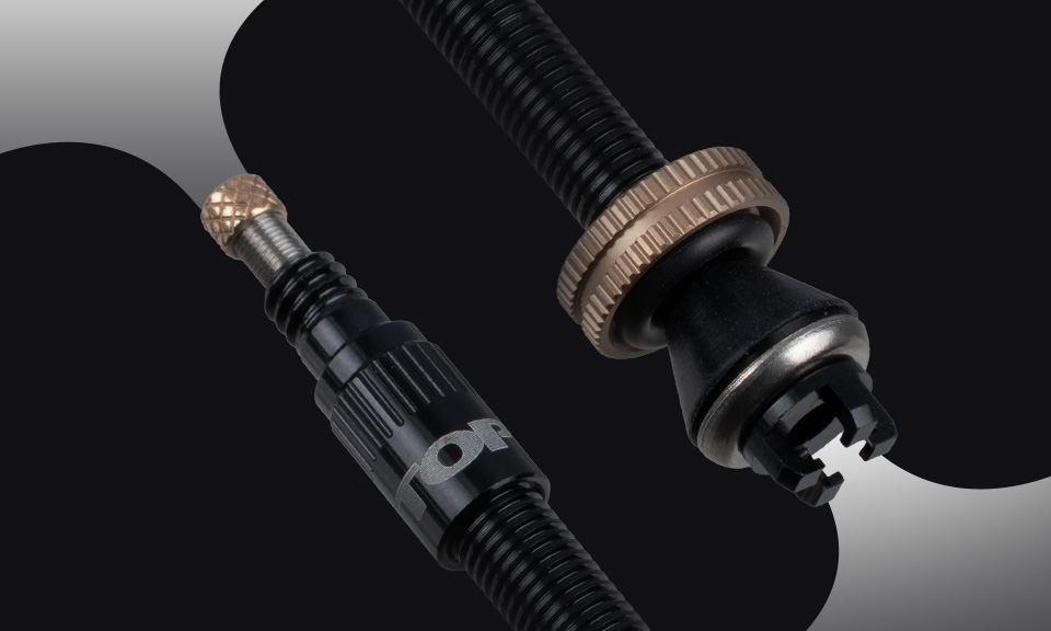 Nahaufnahme eines schwarzen Tubeless-Ventils mit goldfarbener Rändelmutter und Ventilkappe, vor einem abstrakten schwarzen Hintergrund.