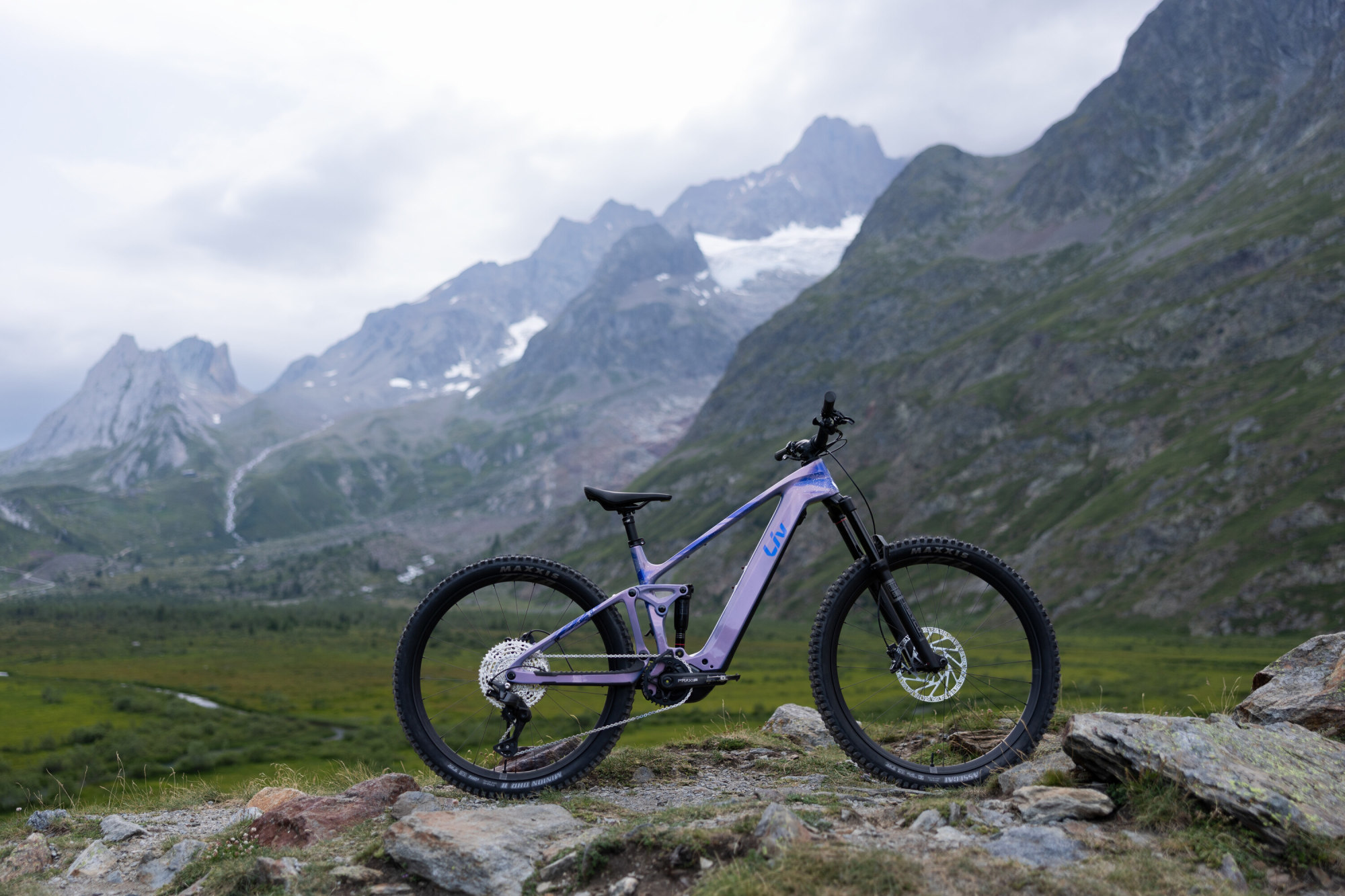Liv Intrigue Advanced E+ 2 E-Mountainbike in Ember Violet vor Alpenkulisse in Seitenansicht.