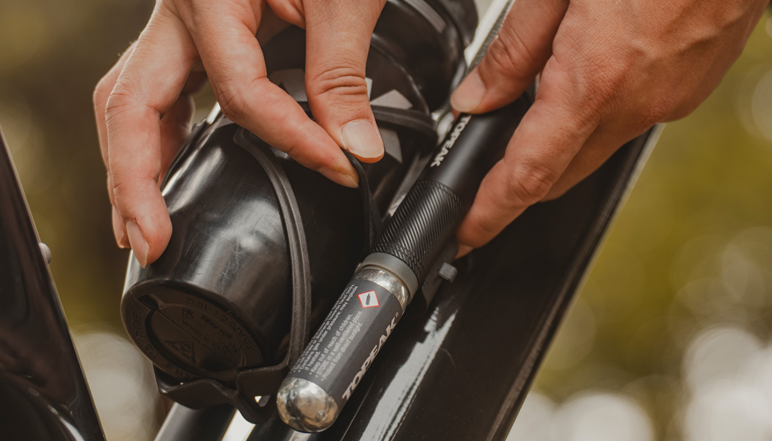  Hände befestigen eine Topeak-Minipumpe mit CO₂-Kartusche an einem Fahrradrahmen unterhalb des Flaschenhalters.